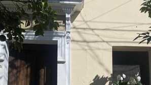 L’entrée d’un immeuble, avec une porte en bois, une petite fenêtre ornée de fleurs blanches et l’ombre projetée par un arbre.