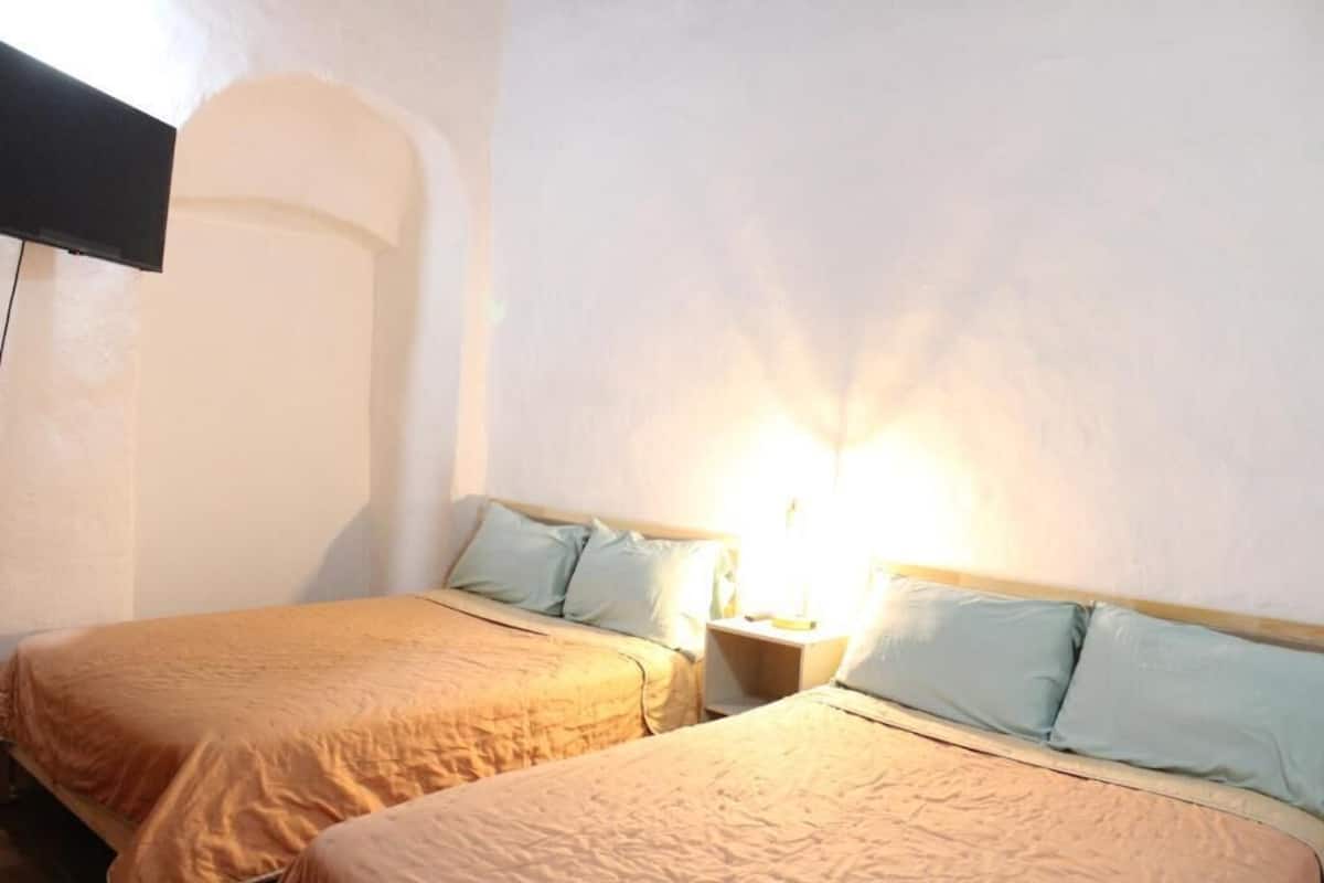 A room with two beds, a nightstand with a lamp, and a TV mounted on the wall.