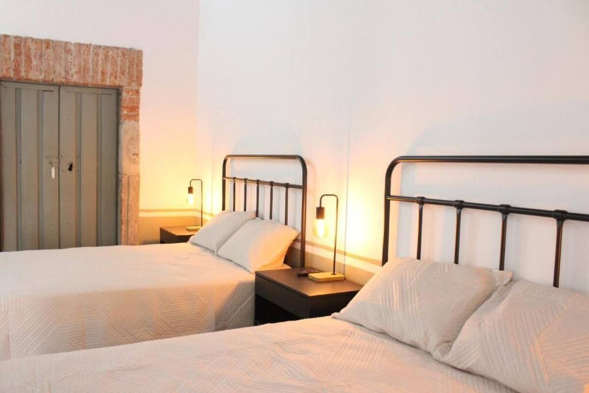 A hotel room with two single beds, a nightstand with a lamp, a wardrobe, and a brick wall.