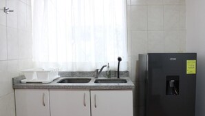 Une cuisine avec des meubles blancs, un réfrigérateur noir, une fenêtre avec des rideaux légers et un plan de travail muni d’un égouttoir et d’un robinet.