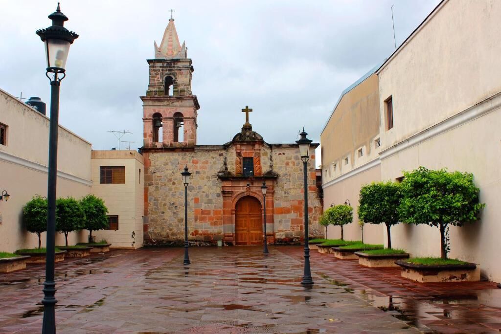 A historic church with a bell tower, a courtyard with a wet pavement, and lamp posts.