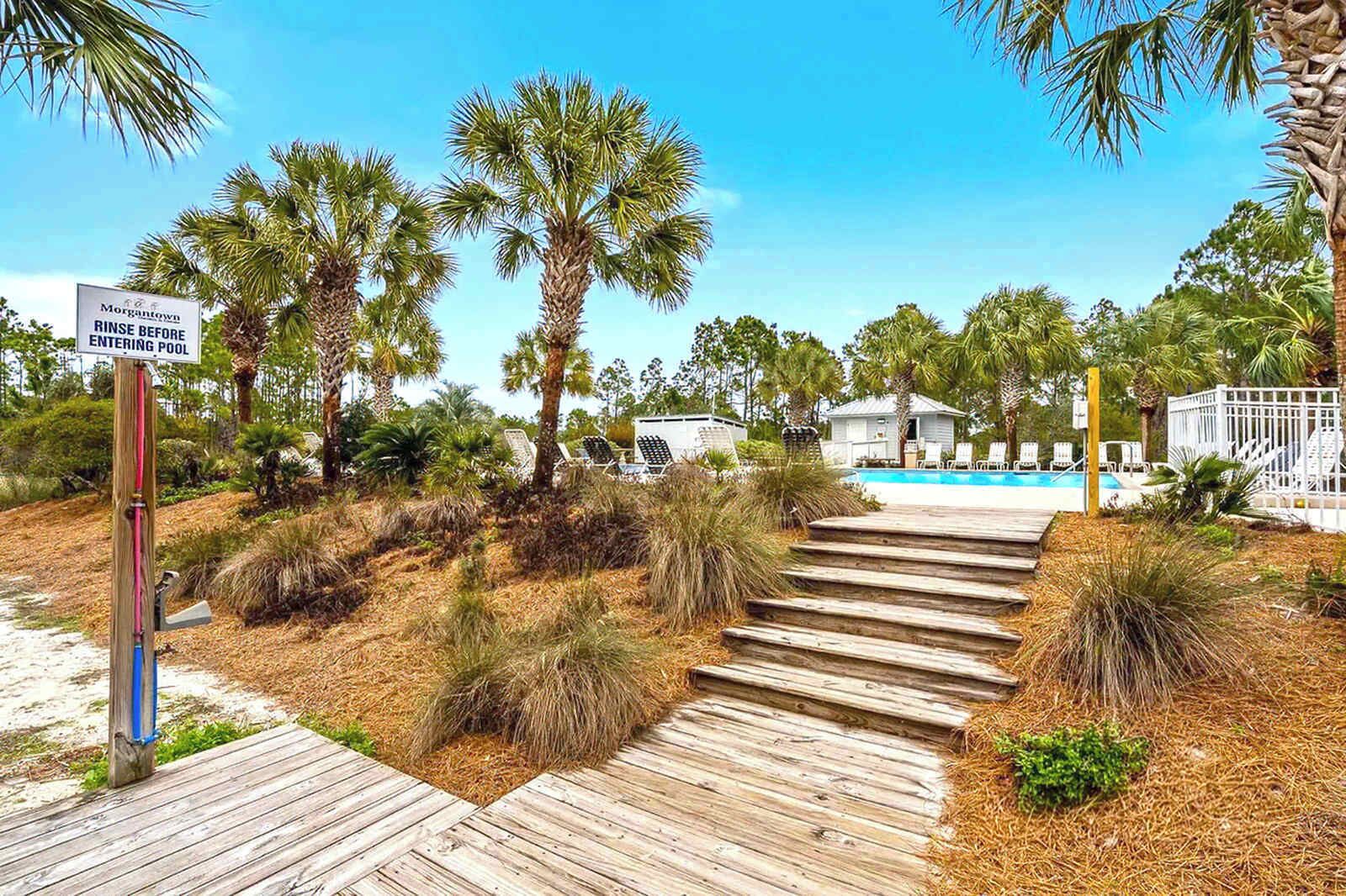 A wooden pathway leading to a pool area with palm trees and a sign indicating to rinse before entering.