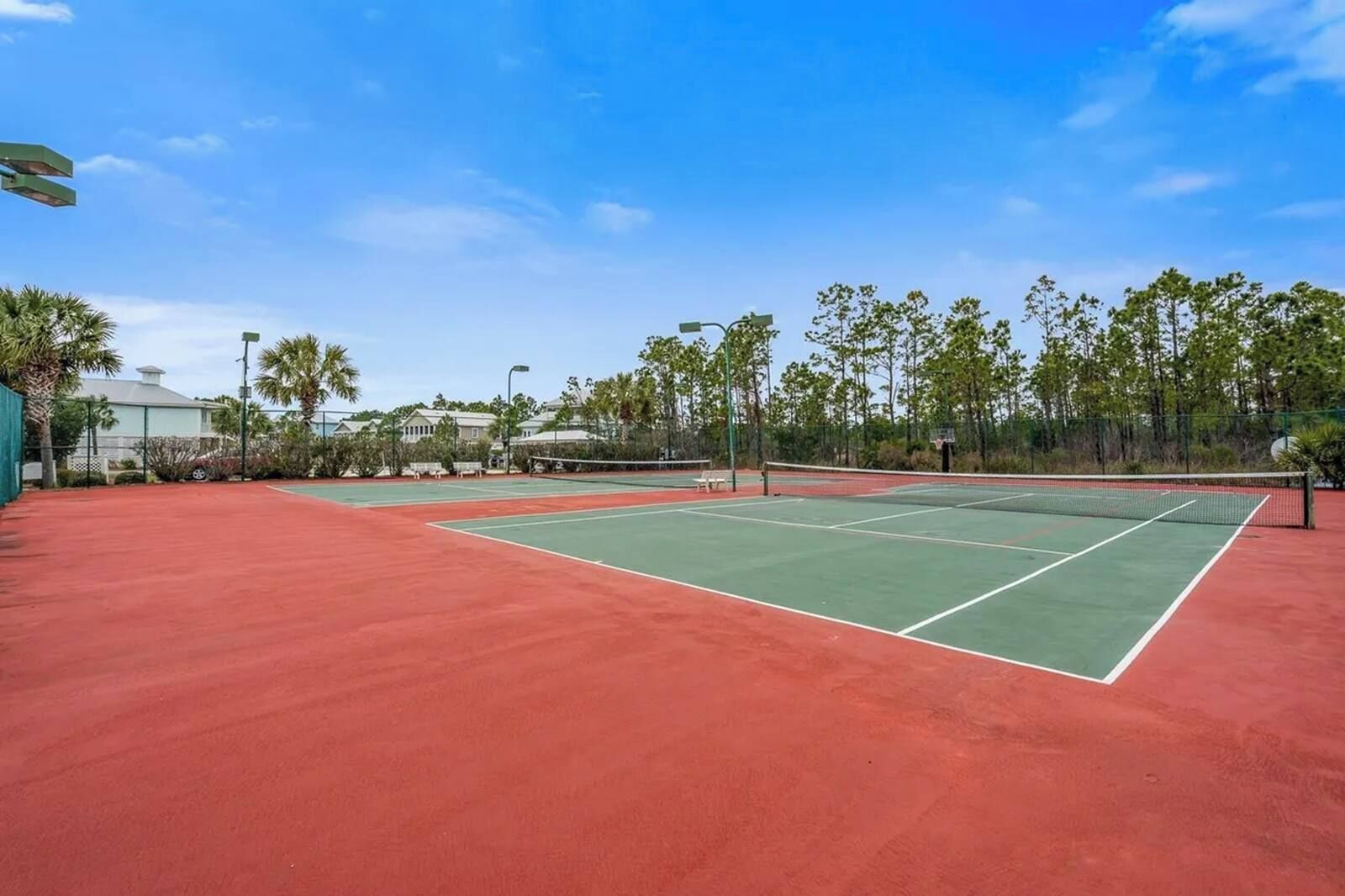 A tennis court with a red surface and a net, surrounded by trees and a building in the background.