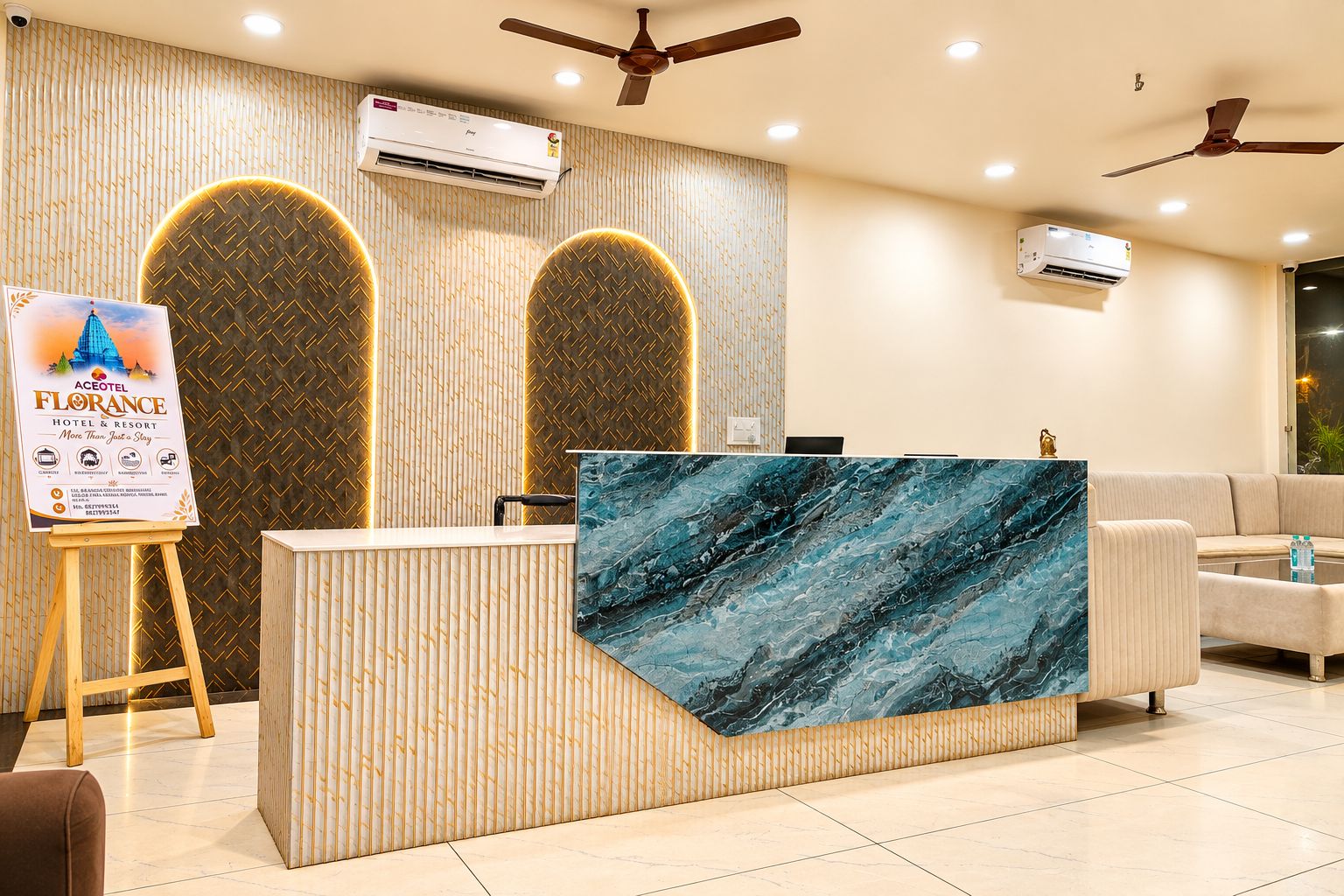 Zona de recepción de hotel con un mostrador de mármol, dos unidades de aire acondicionado, un cartel con marco de madera y un ventilador de techo.