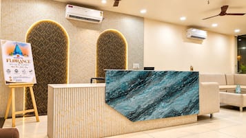 Zona de recepción de hotel con un mostrador de mármol, dos unidades de aire acondicionado, un cartel con marco de madera y un ventilador de techo.