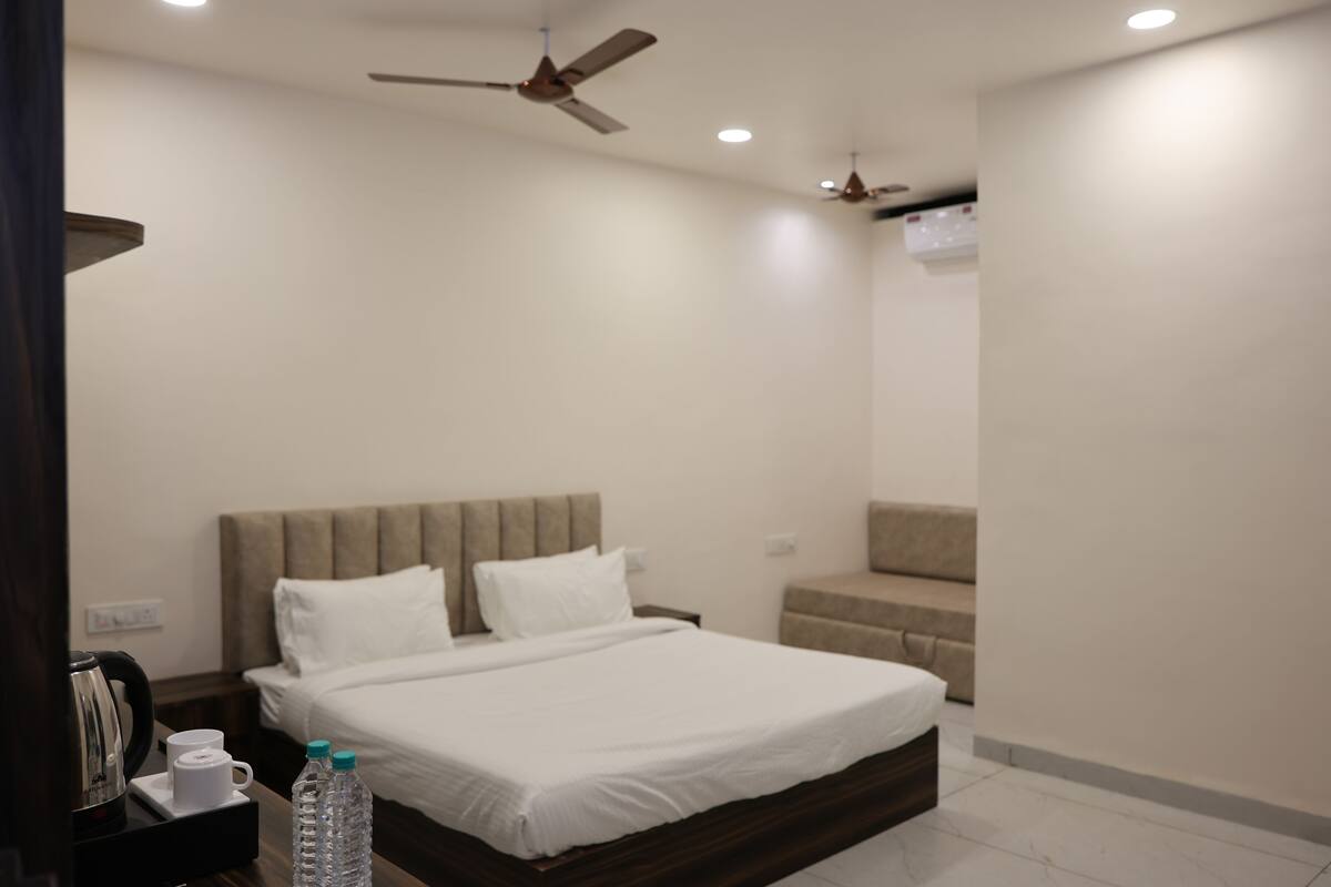 A hotel room with a bed, bedside tables, a sofa, ceiling fans, and an air conditioning unit.