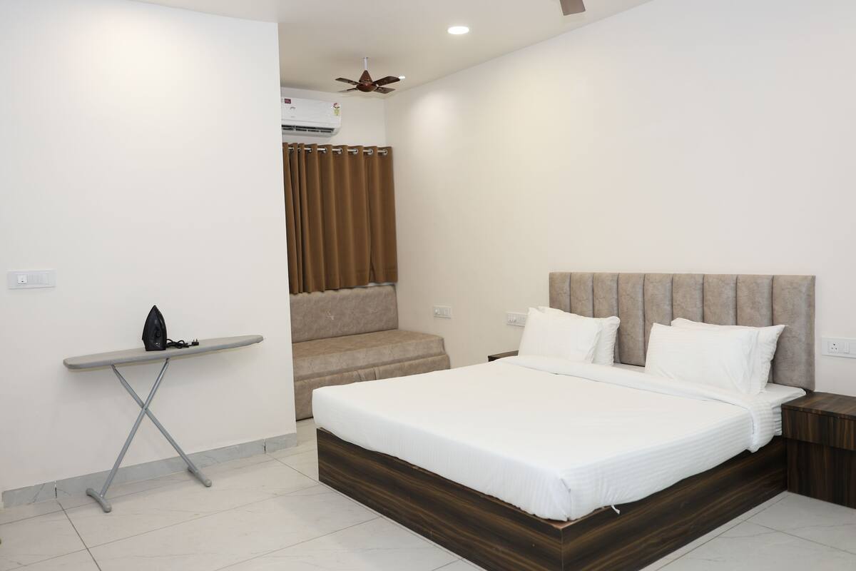 A bedroom with a bed, a small table with an iron, a ceiling fan, and a window with curtains.