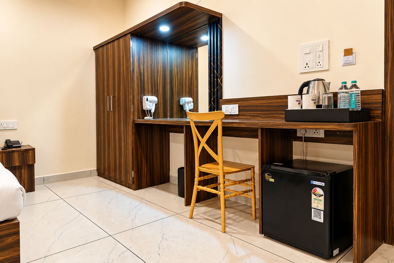A compact hotel room with a wooden wardrobe, a small desk with a chair, a mini fridge, and a coffee machine.