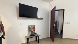 A room with a flat-screen TV mounted on the wall, a small table with a painting, a chair, and a door leading to another room. - Delightful 1-bedroom apartment with AC in Sosua (Sosua)