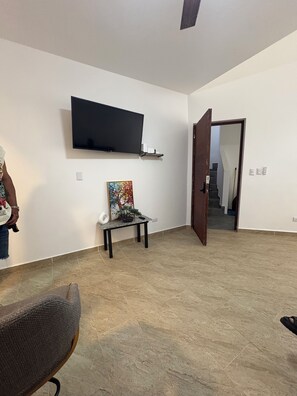 Ein Zimmer mit einem an der Wand befestigten Flachbildfernseher, einem kleinen Tisch mit einem Gemälde, einem Stuhl und einer Tür zu einem weiteren Raum.