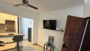 A modern kitchen with granite countertops, a dining area with a dark chair, a TV mounted on the wall, and a wooden door. - Delightful 1-bedroom apartment with AC in Sosua (Sosua)
