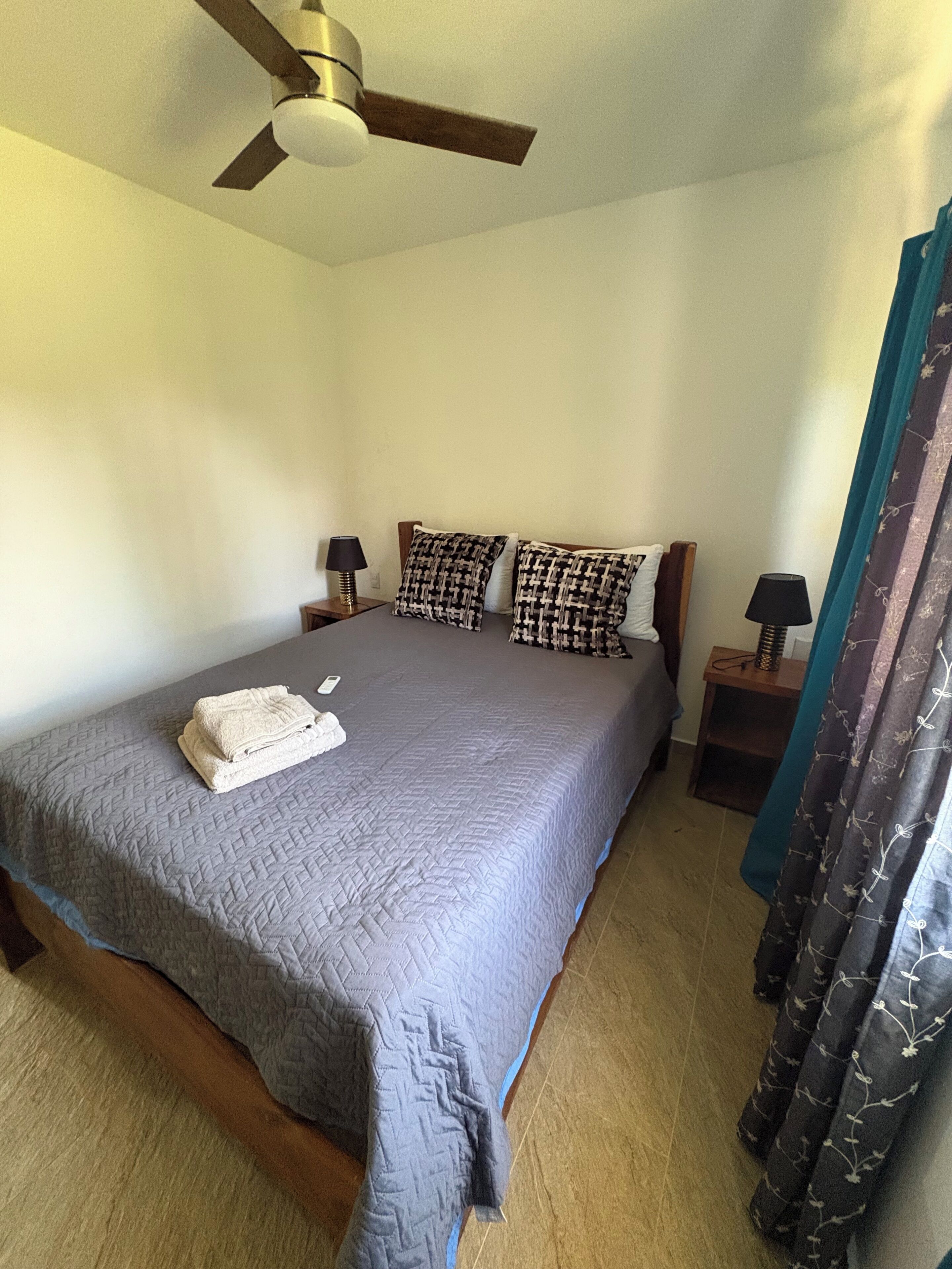 A bedroom with a bed, a ceiling fan, two bedside tables with lamps, and a curtain.
