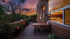 Um pátio com mesa e cadeiras, vista para uma cozinha através de uma janela e um pôr do sol ao fundo.
