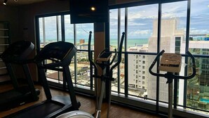 Academia com esteiras, elíptico e bicicleta ergométrica, com vista para a cidade através de grandes janelas.