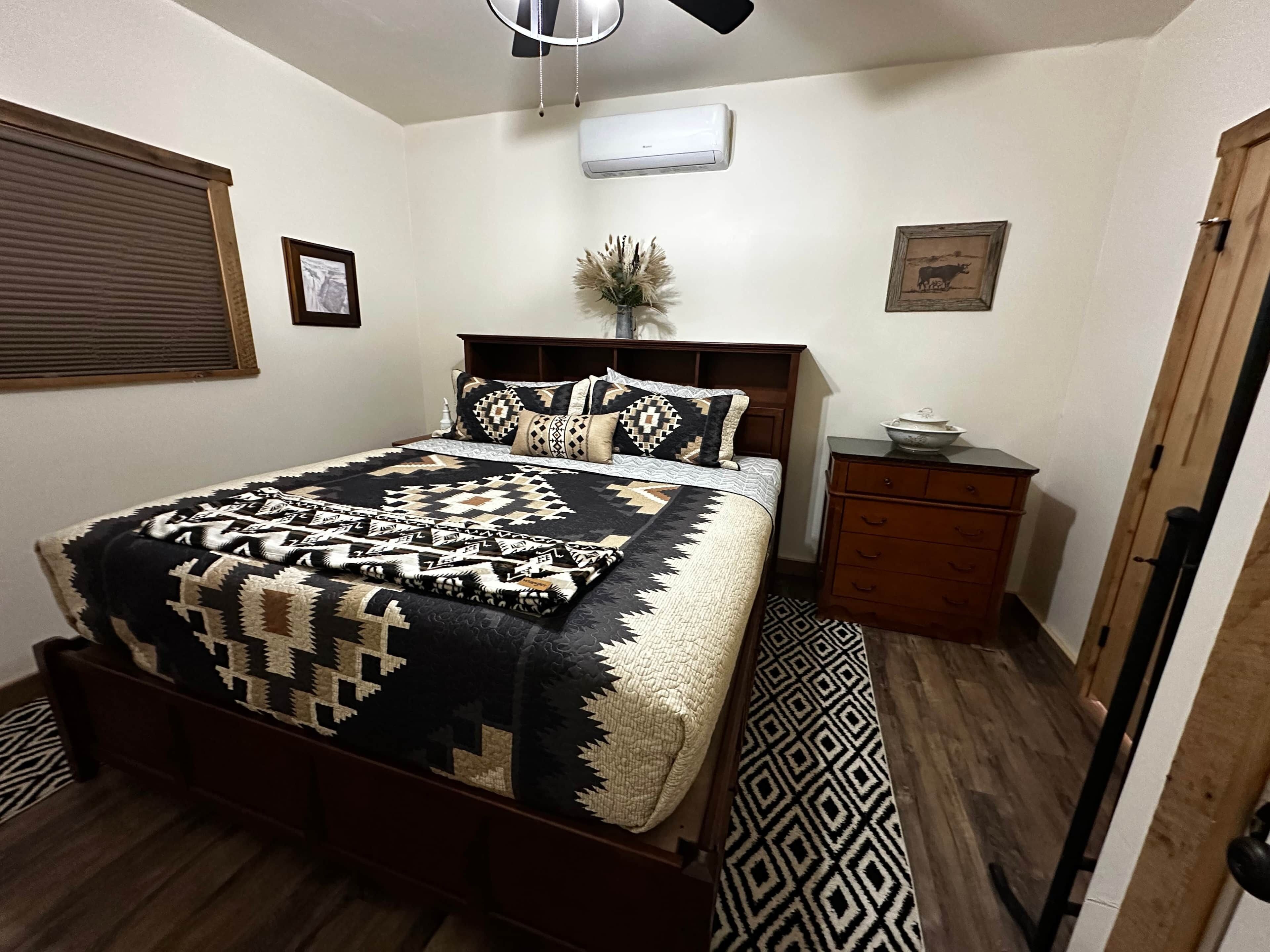 A bedroom with a bed, a nightstand, a dresser, a window with blinds, and an air conditioning unit.