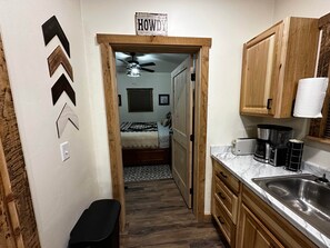 Corredor estreito que dá para um quarto com cama, ventilador de teto e janela com persianas. Há um porta-de-madeira com uma placa acima, uma pia com torneira e armários de madeira.