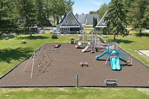 A playground with slides and climbing structures, surrounded by grass and trees, with houses in the background.