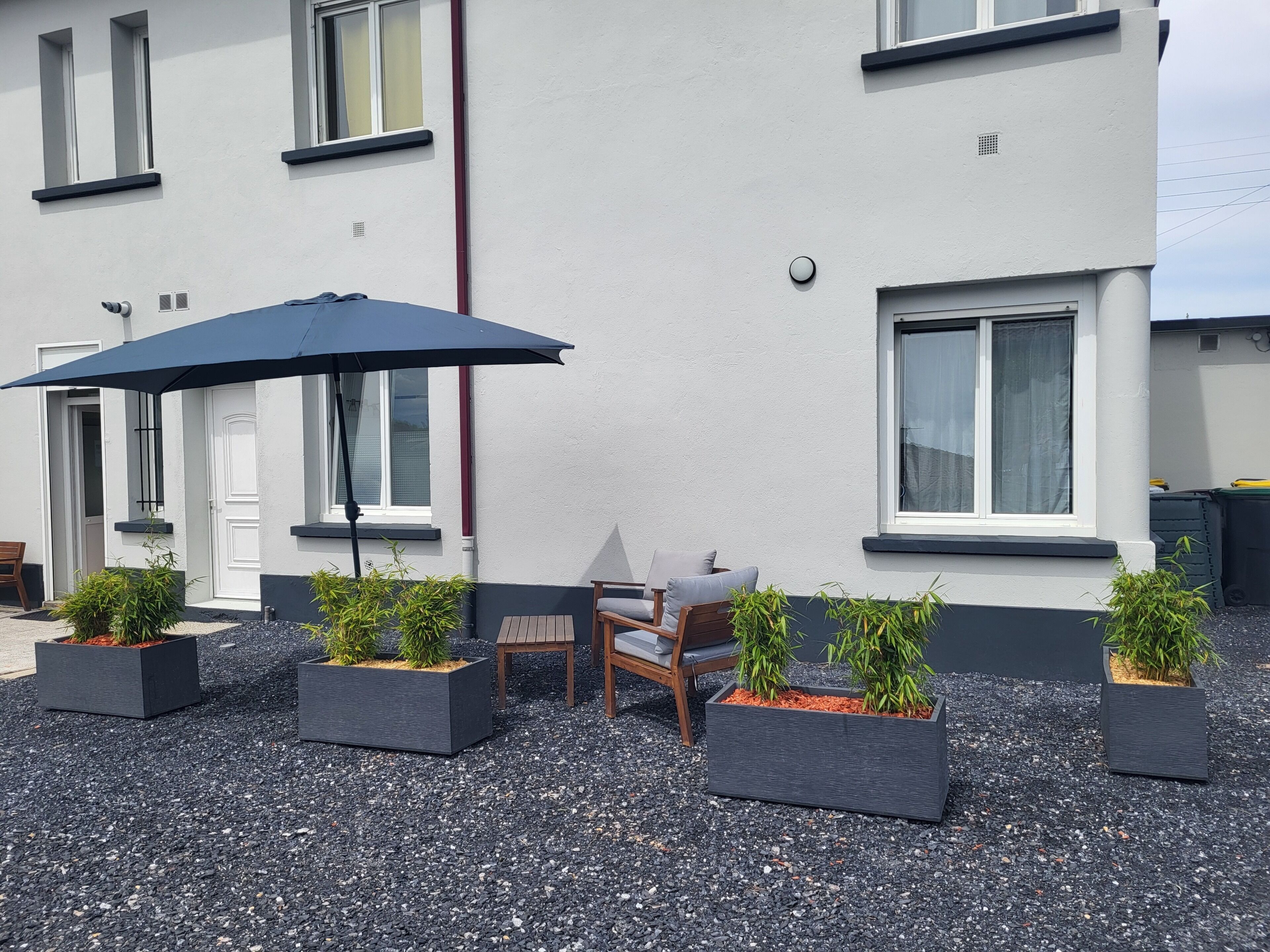 Un bâtiment à l’extérieur blanc, avec des encadrements de fenêtres noirs et une porte. On y trouve des espaces aménagés à l’extérieur avec une table et des chaises, ainsi que plusieurs plantes en pot.