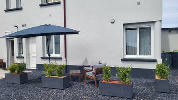 Un bâtiment à l’extérieur blanc, avec des encadrements de fenêtres noirs et une porte. On y trouve des espaces aménagés à l’extérieur avec une table et des chaises, ainsi que plusieurs plantes en pot.