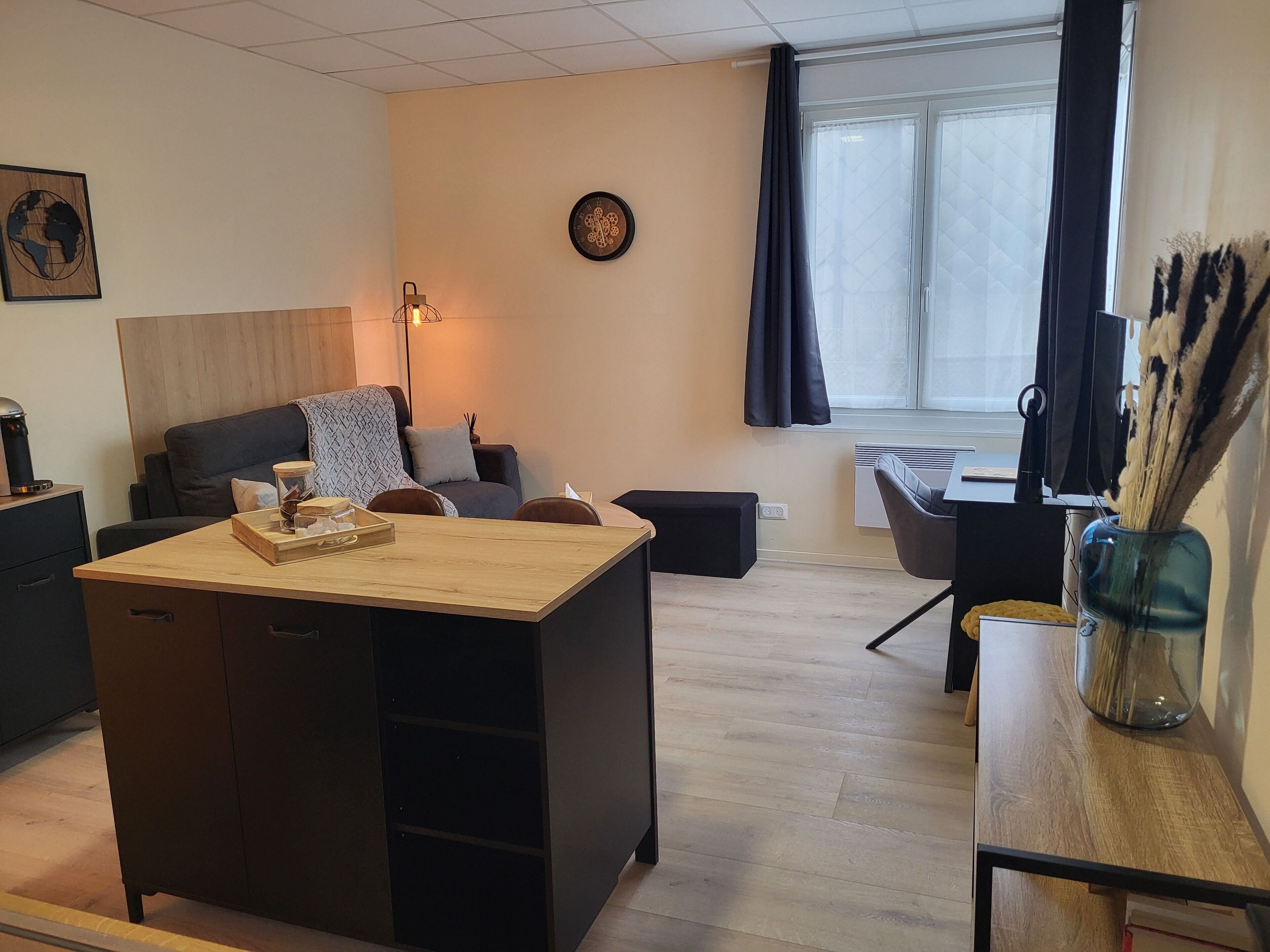 Un salon moderne avec une table basse en bois, un canapé, un bureau et une chaise. Il y a une horloge au mur et une fenêtre avec des rideaux.