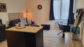 Un salon moderne avec une table basse en bois, un canapé, un bureau et une chaise. Il y a une horloge au mur et une fenêtre avec des rideaux.
