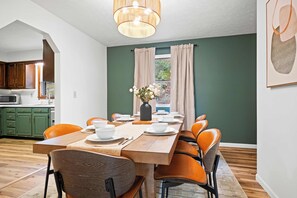 Ein Essbereich mit einem Holztisch, orangefarbenen Stühlen, einer Vase mit Blumen und Blick auf eine grüne Außenansicht durch ein Fenster.