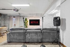 Ein modernes Wohnzimmer mit einem grauen Ecksofa, einem Wand-Fernseher und einem großen Lautsprecher auf einem Stativ.