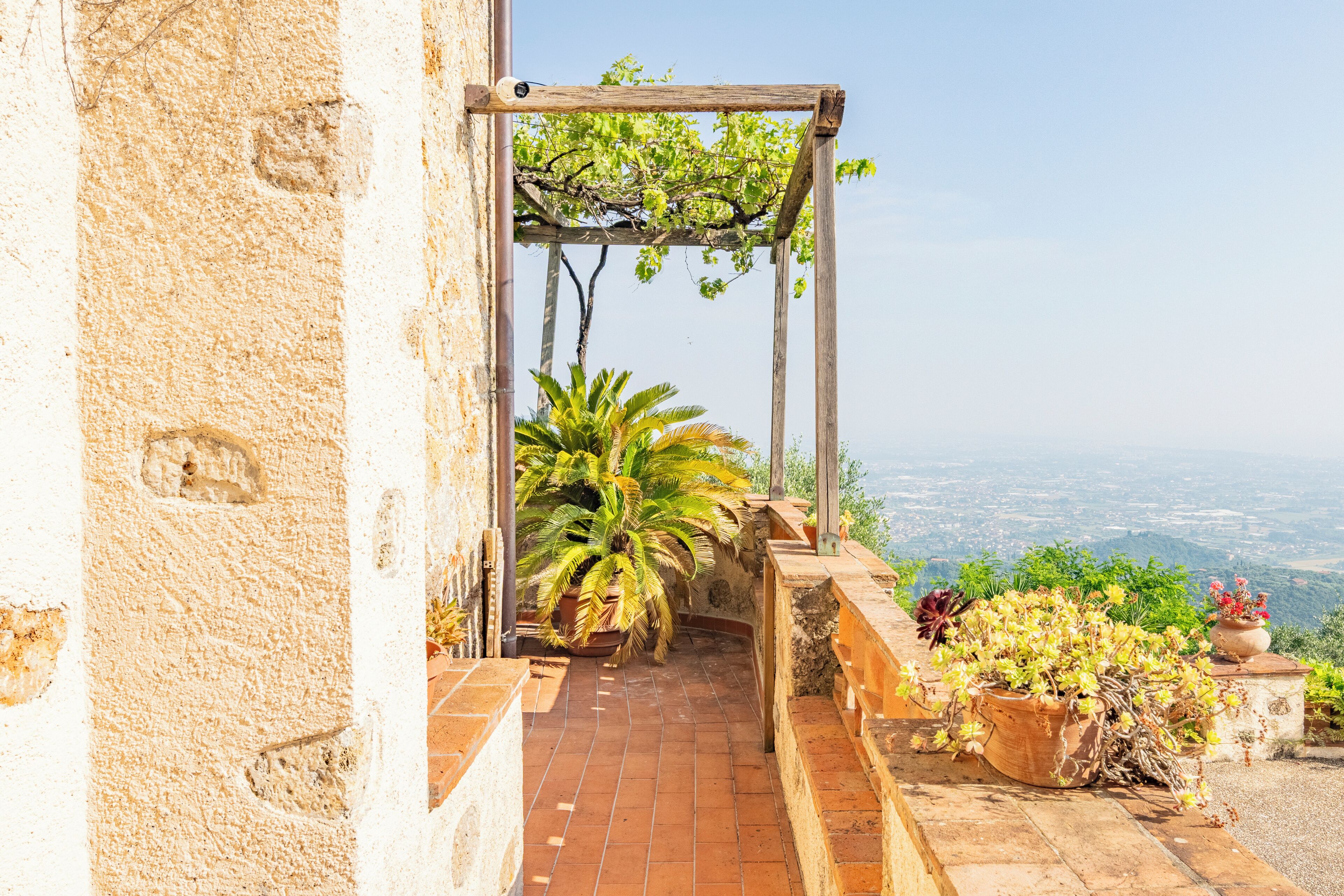 Ein steiniger Weg führt zu einer Holzpergola mit Pflanzen, mit Blick auf eine Stadtlandschaft.