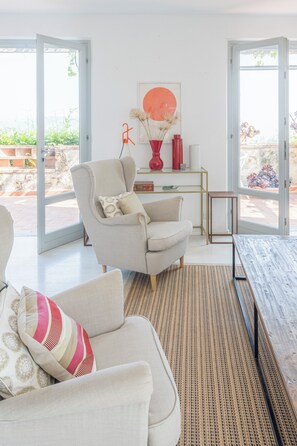 Ein offener Wohnbereich mit Sofa, Sessel und Couchtisch, einem Schreibtisch mit Vase und Bild, sowie großen Glasüren, die zu einem Außenbereich führen.
