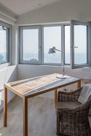 Ein Zimmer mit Holztisch, Korbstuhl und Meerblick durch große Fenster.