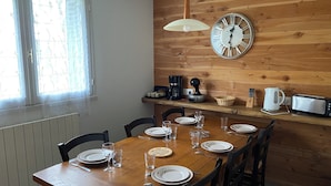 A dining area with a wooden table set for six, a wall clock, a coffee maker, a toaster, and a window with floral curtains.