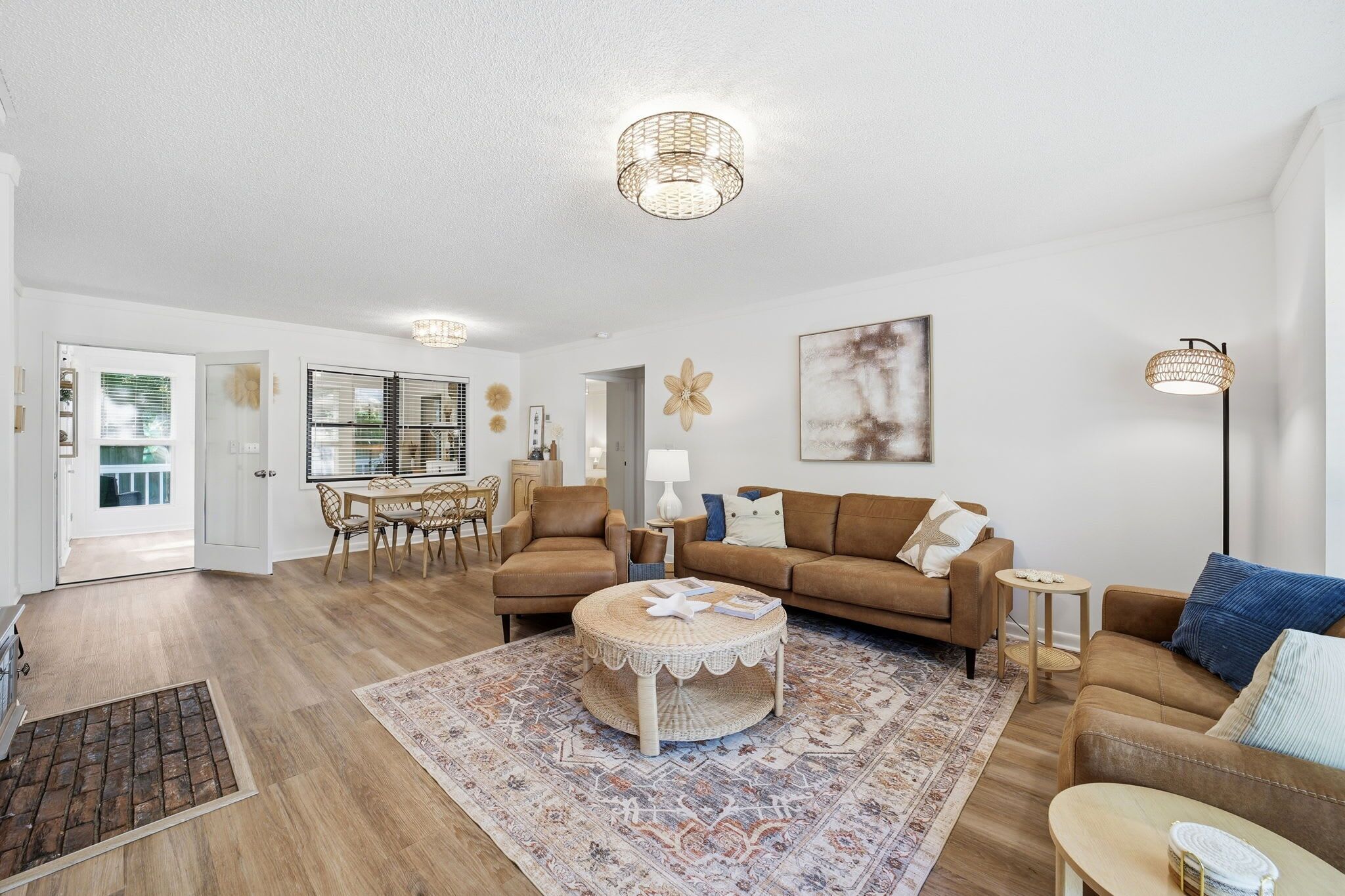 A spacious living room with a round coffee table, two sofas, a dining area with a table and chairs, and a large window with blinds.