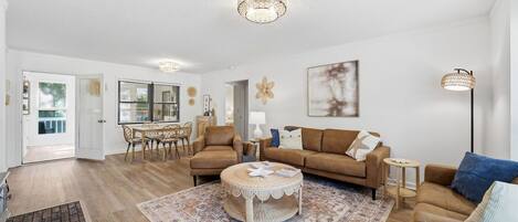 A spacious living room with a round coffee table, two sofas, a dining area with a table and chairs, and a large window with blinds.