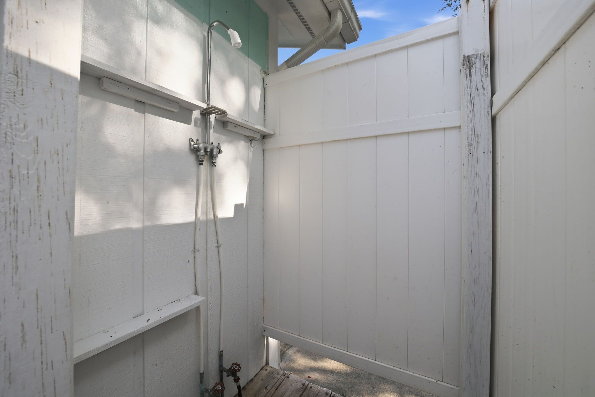 A white wooden structure with a metal shower fixture and a small step leading to a door.
