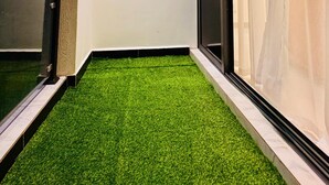 Een balkon met kunstgras, een raam met dunne gordijnen en een rond spiegel aan de muur.
