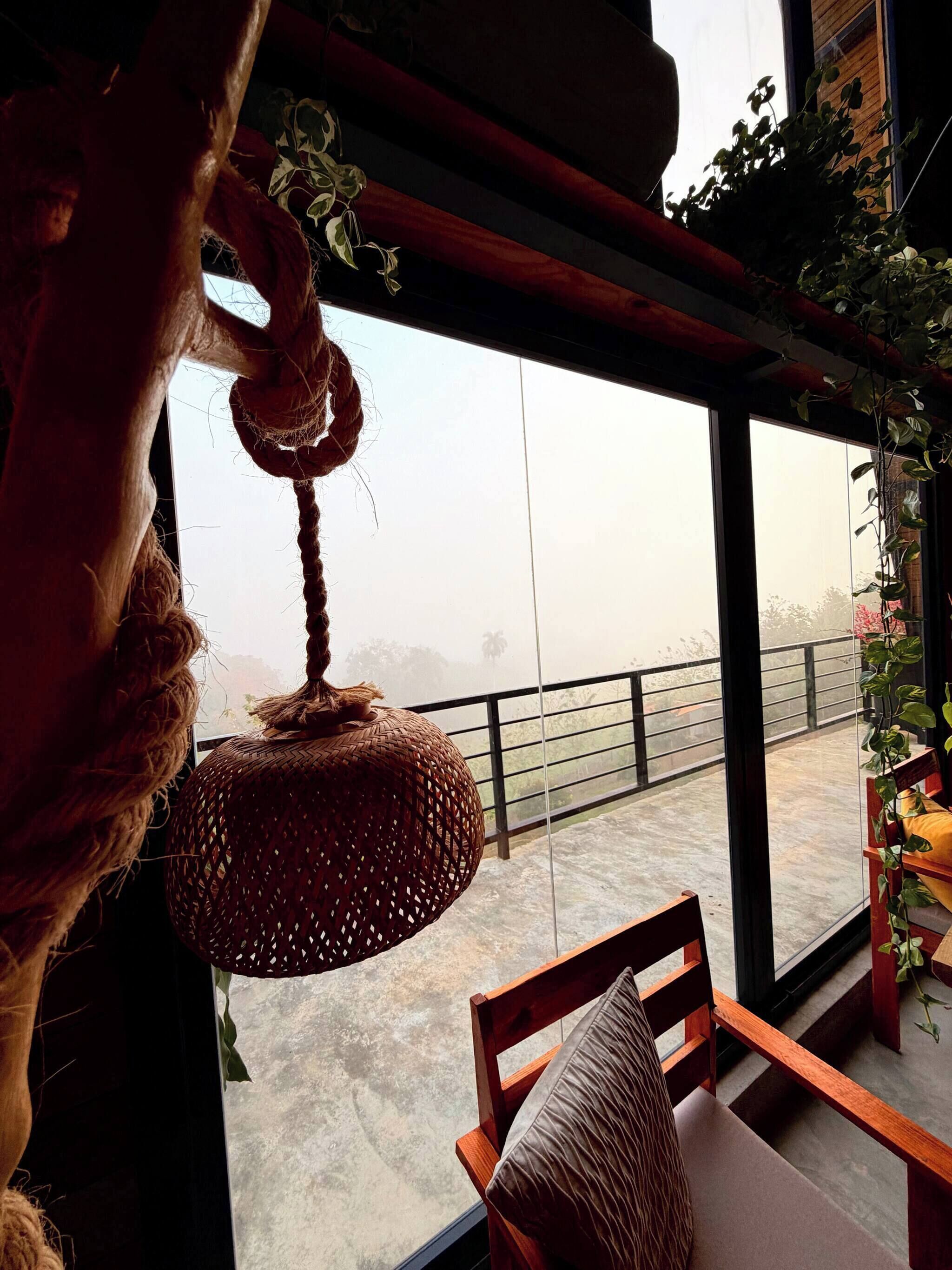 A room with a large window, a hanging woven light fixture, a wooden chair, and a view of a balcony with railings.