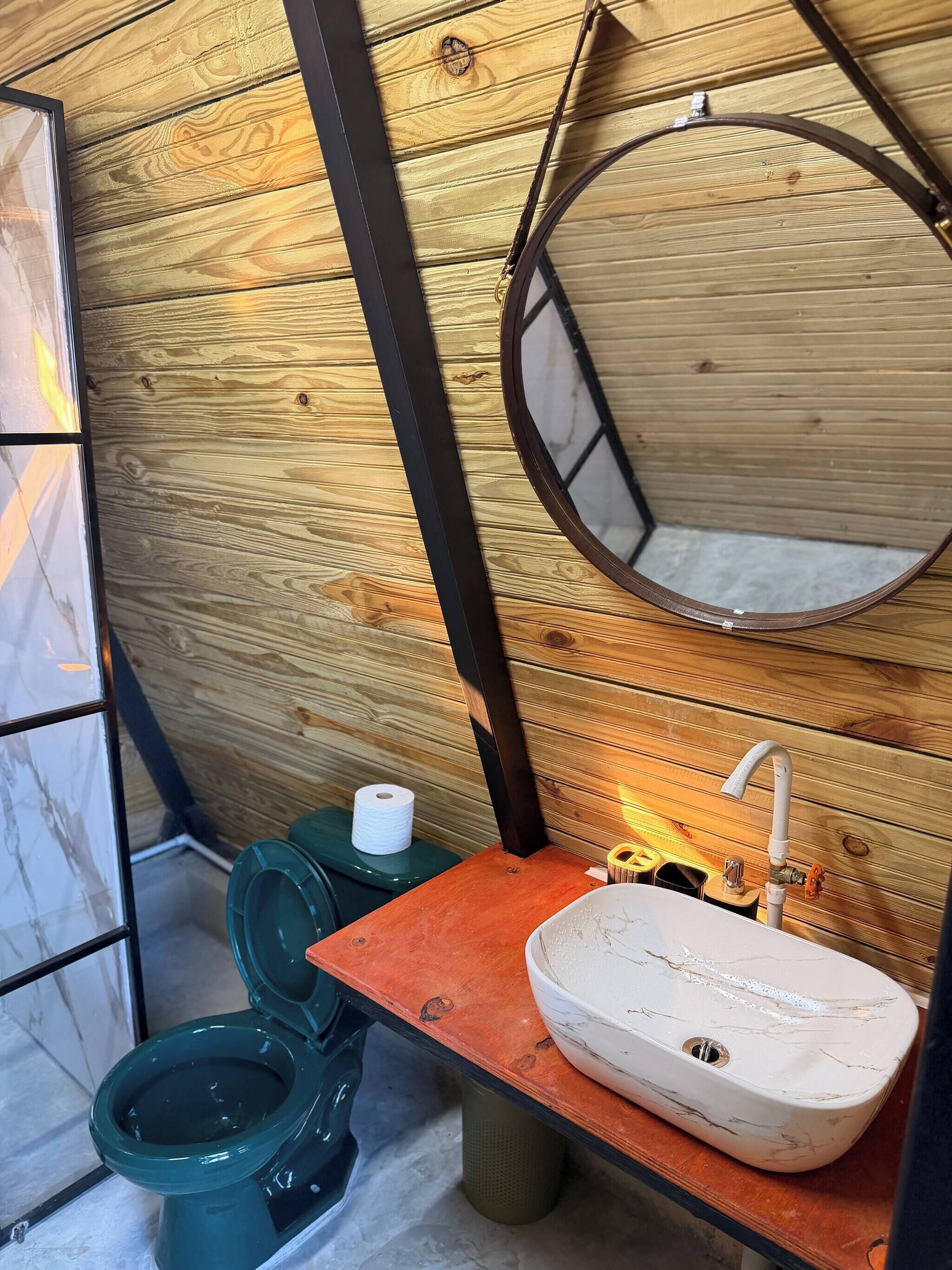 Un baño pequeño con detalles de madera, un inodoro verde, un lavamanos blanco con diseño similar al mármol y un espejo en la pared.