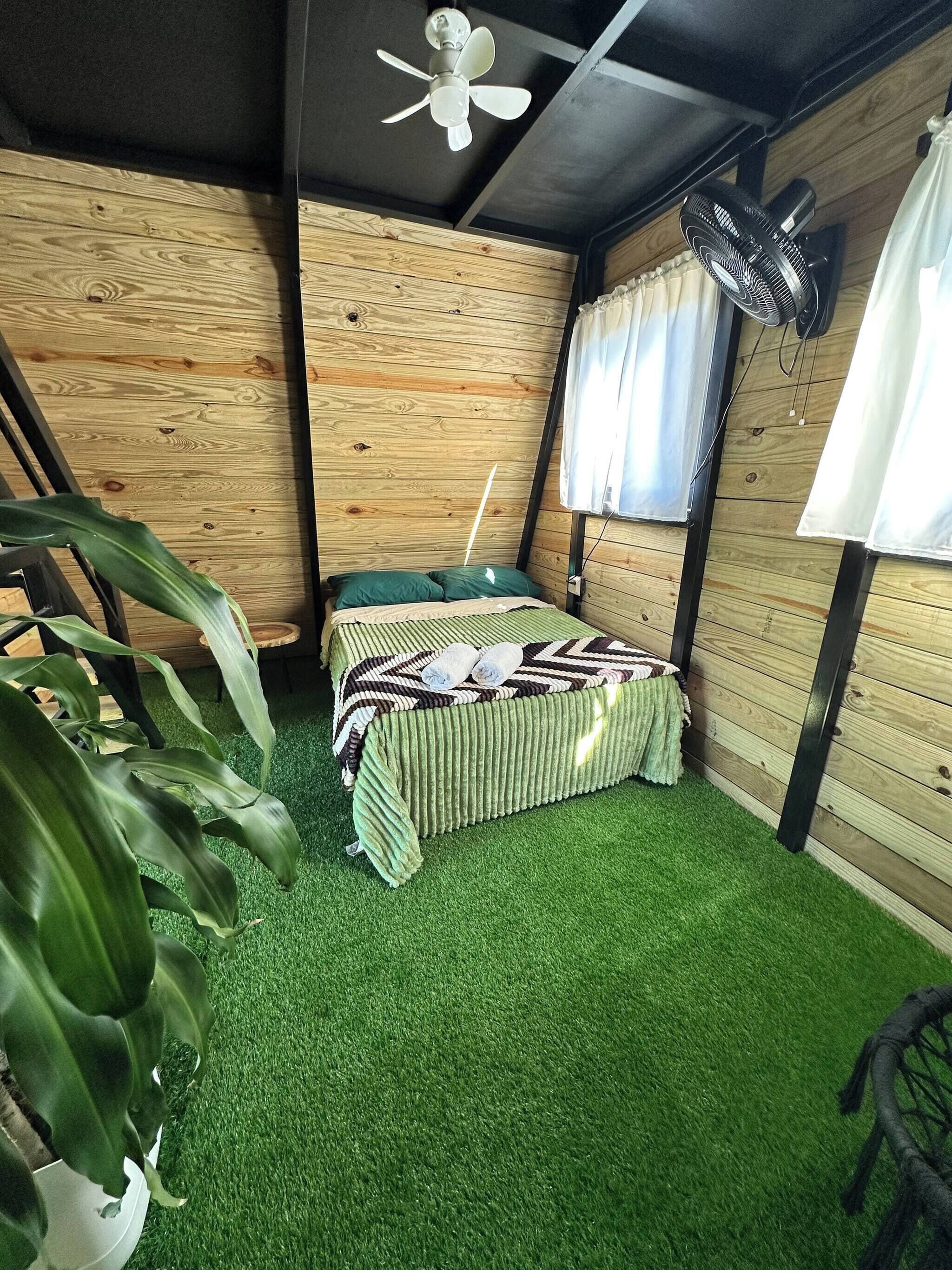 A room with a bed, artificial grass floor, wooden walls, a ceiling fan, and a window with curtains.