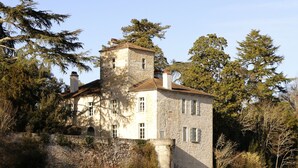 A stone building with a tower, situated by a calm body of water, surrounded by trees. - À l'ombre du grand pin - Bed and breakfast on the banks of the Lot and artist's studio! (Saint-Sylvestre-sur-Lot)