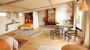 A spacious room with a bed, a dining area with chairs, a round table, and a large window with curtains. - À l'ombre du grand pin - Bed and breakfast on the banks of the Lot and artist's studio! (Saint-Sylvestre-sur-Lot)
