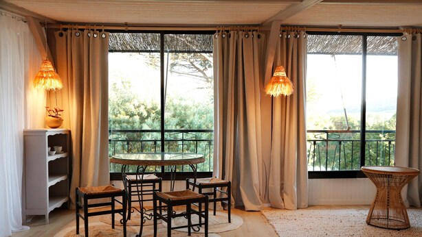 A room with a round table, four chairs, a small side table, a shelf with dishes, and a view through large windows. - À l'ombre du grand pin - Bed and breakfast on the banks of the Lot and artist's studio! (Saint-Sylvestre-sur-Lot)