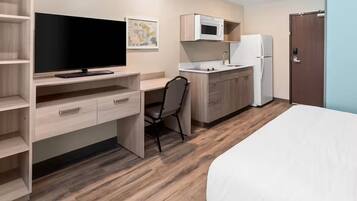 Une chambre d’hôtel compacte comprenant un lit, un bureau avec un téléviseur à écran plat, une chaise, une kitchenette équipée d’un four à micro-ondes et d’un réfrigérateur, ainsi qu’une porte.