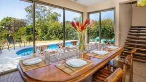 Comedor con vista a la piscina, una mesa de madera preparada para cuatro personas y una escalera a la derecha.