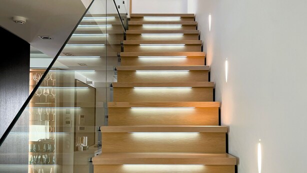 Una escalera moderna con escalones de madera y barandillas de vidrio, que conduce a un espacio con una vitrina de copas de vino.