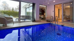 Una piscina interior moderna con azulejos azules y cristalinos, un área de patio con techo de vidrio y asientos, y vistas al exterior.