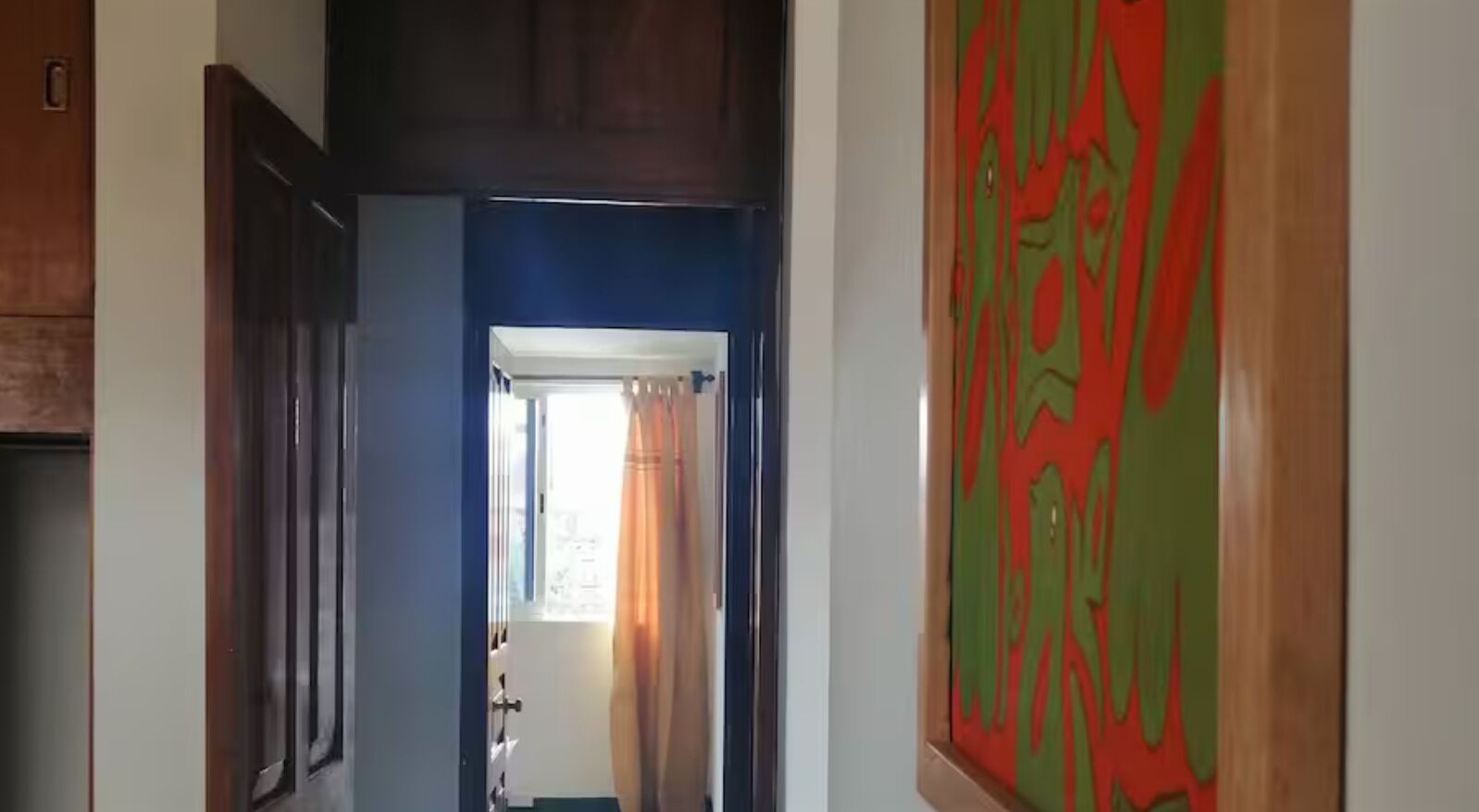 Un corridoio con una porta che dà su una stanza, una finestra con tende e un quadro astratto dai colori vivaci appeso al muro.
