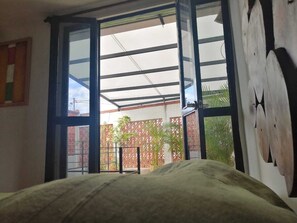 Ein Zimmer mit einem Bett, einem großen Fenster und Blick auf einen Außenbereich mit Pflanzen und einem Zaun.