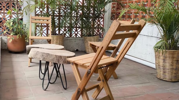 Un patio exterior con mobiliario de madera, incluyendo una mesa y sillas, rodeado de plantas en macetas y una valla decorativa.