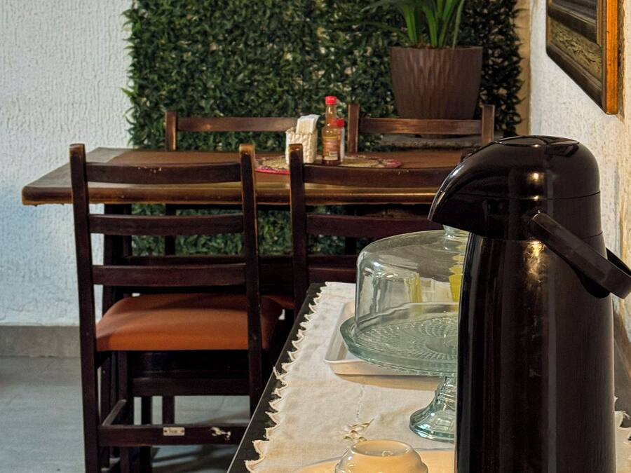 Un comedor con una mesa dispuesta para dos personas, que incluye una cafetera, un expositor de vidrio para pasteles y una planta en maceta.