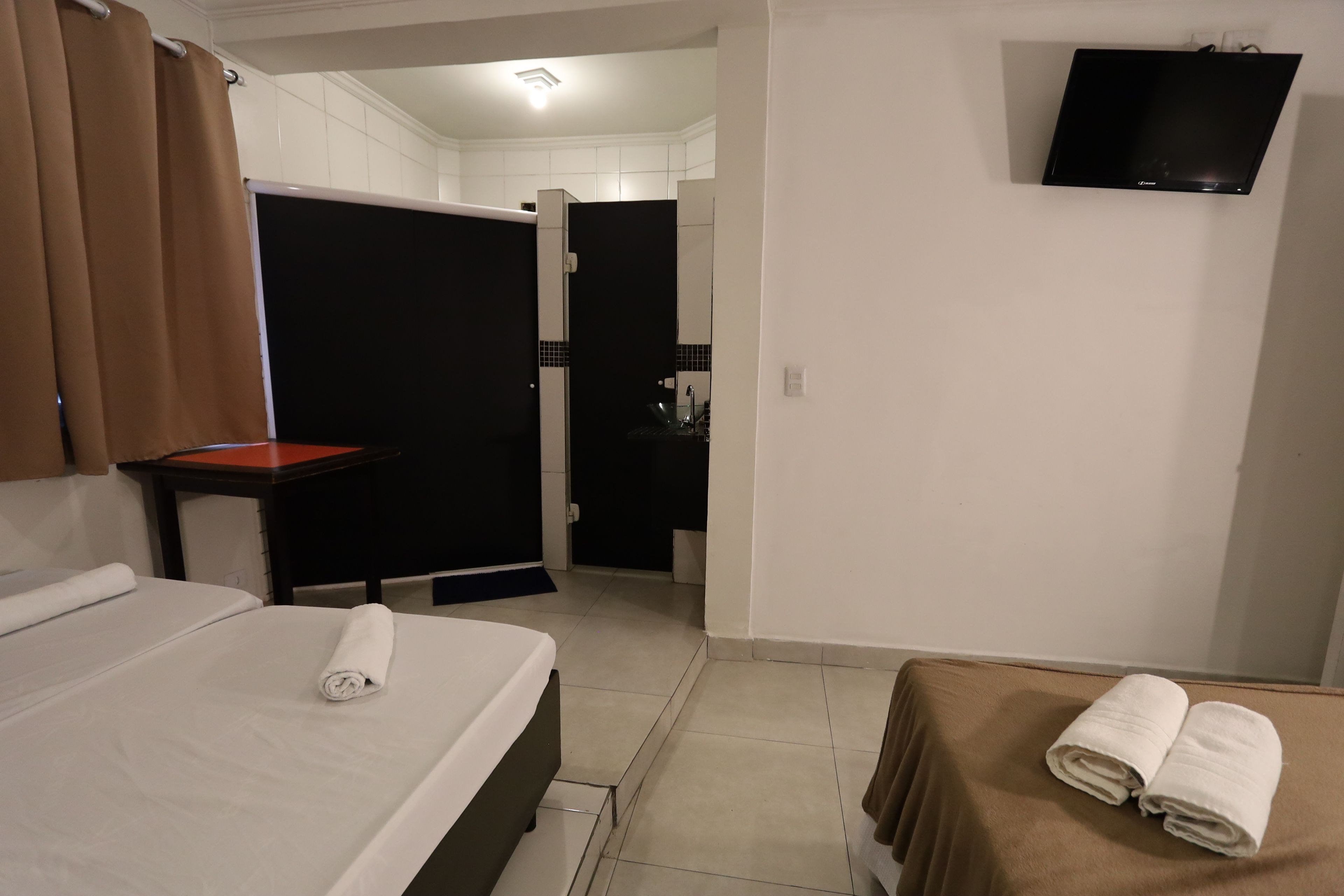 Une chambre d’hôtel avec un lit, une table, un téléviseur fixé au mur et une salle de bain visible à travers une porte ouverte.